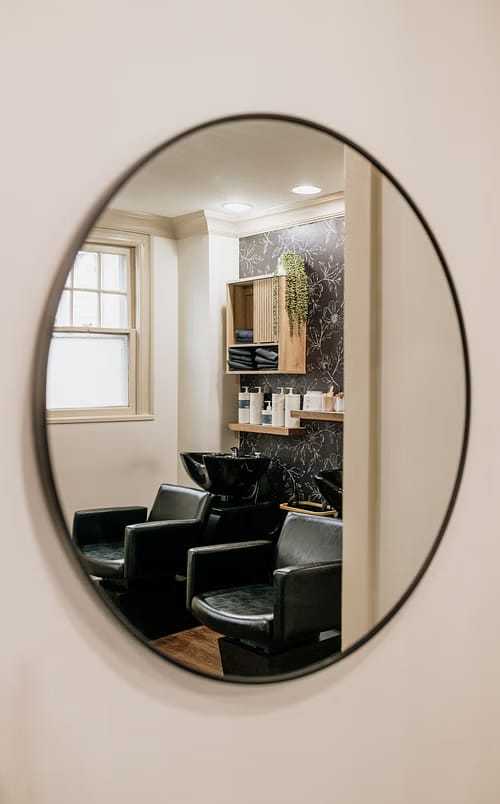 Reflection of a modern hair salon with styling chairs and a washbasin in an oval mirror.