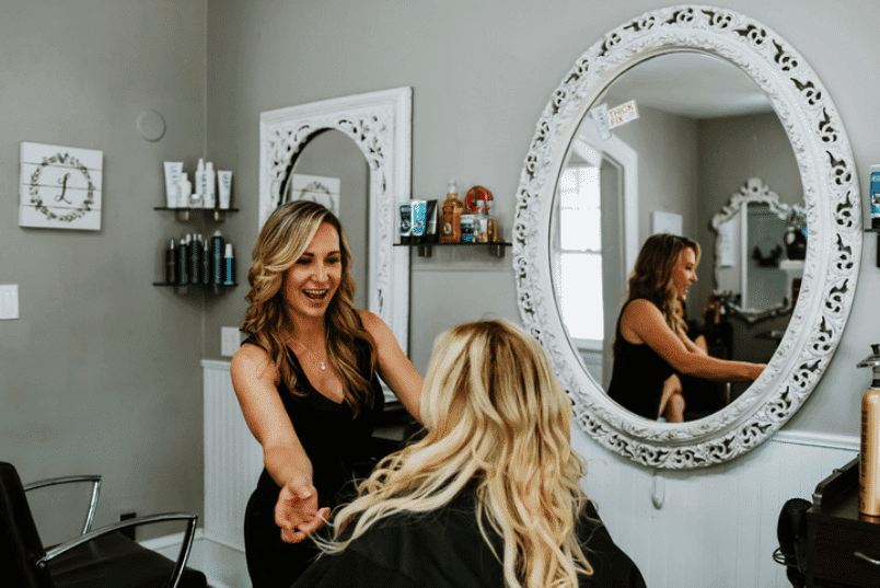A hairstylist laughing while styling a client's hair in a chic, modern salon.